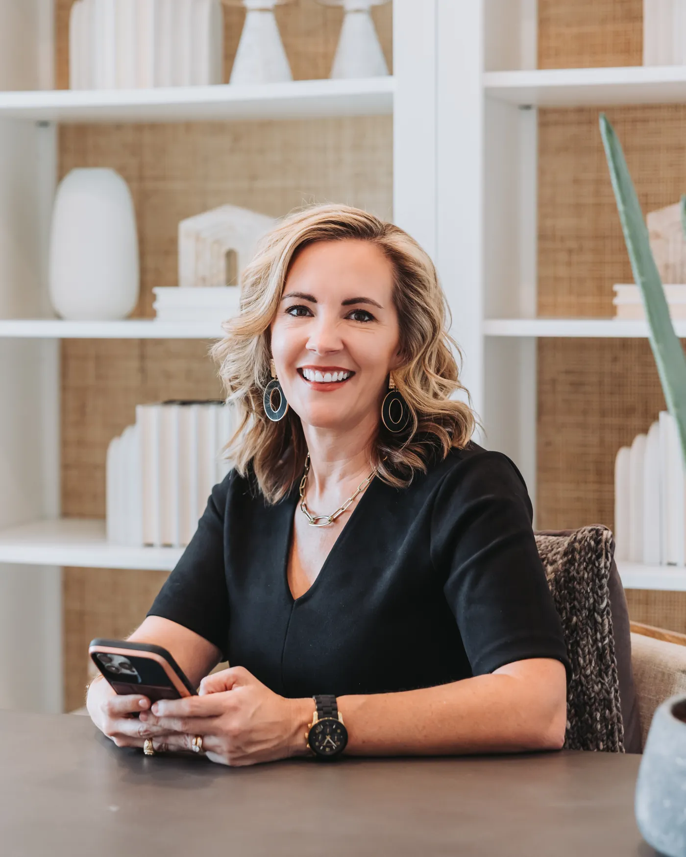 Lucy Thornton seated at a table holding her phone, smiling at the camera, with a white bookshelf behind her