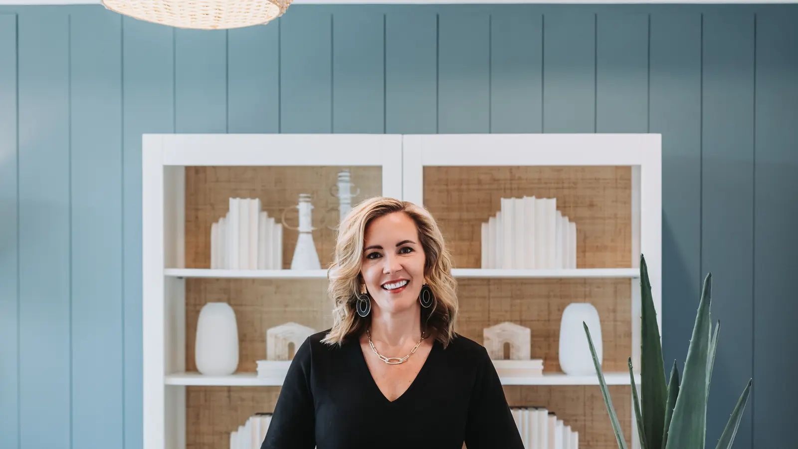Lucy Thornton sitting on a desk under a woven pendant light in a blue-panelled office