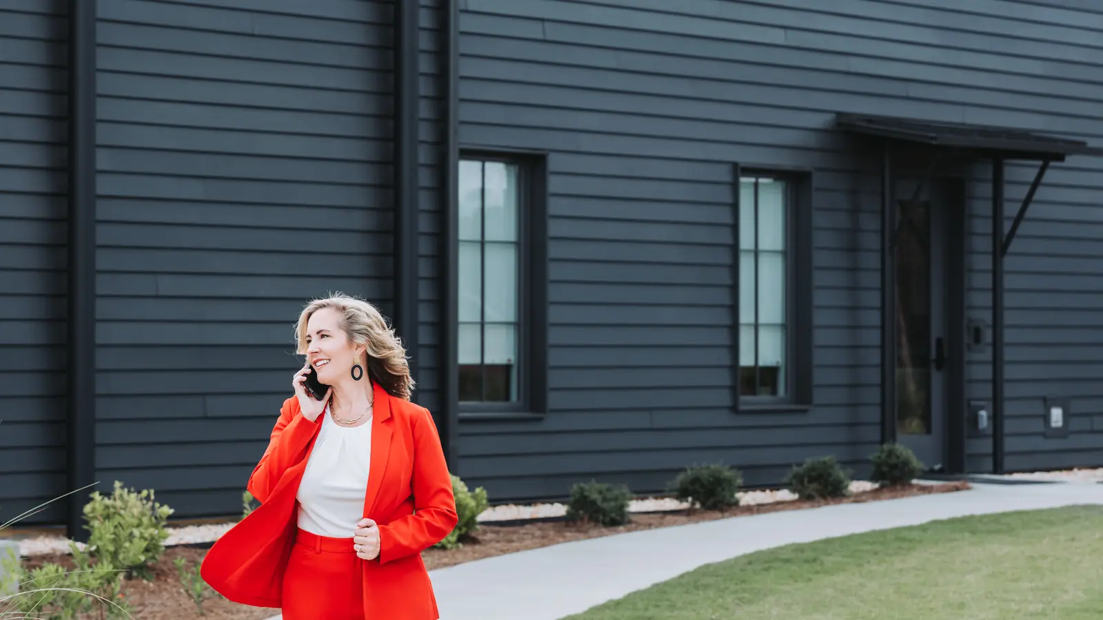 Lucy Thornton in a red suit walking along a path beside a black modern building